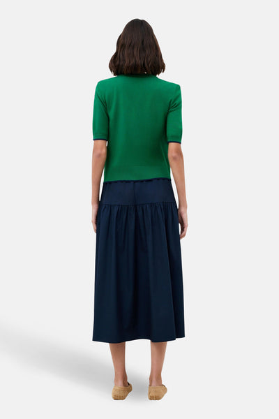 Cardigan vert émeraude par CAROLL, manches courtes, porté avec une jupe midi bleu marine plissée.
