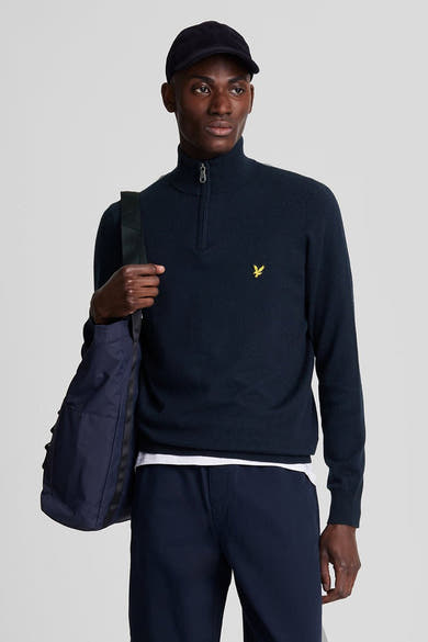 Pull à col roulé bleu de LYLE & SCOTT, porté avec un pantalon bleu marine et une casquette.