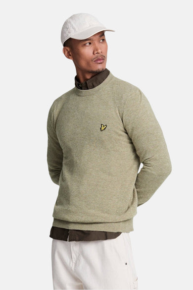 Pull à col rond vert de LYLE & SCOTT, porté sur une chemise marron et un pantalon blanc, avec une casquette blanche.
