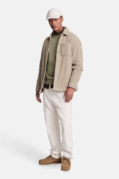 Pull à col rond vert de LYLE & SCOTT, porté avec une chemise beige et un pantalon blanc.