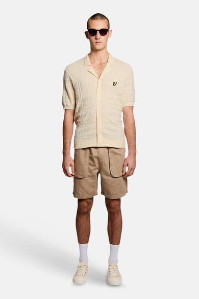 Ecru gilet van LYLE & SCOTT met getextureerde cubaanse kraag en geel adelaarslogo, gedragen met een beige short, lichte sneakers en een zonnebril.