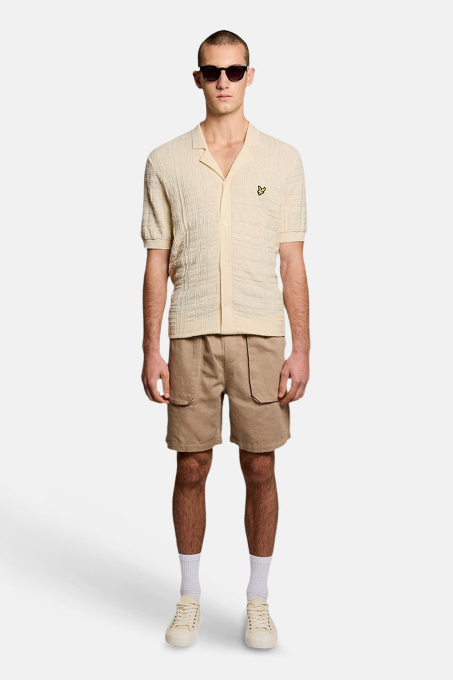 Ecru gilet van LYLE & SCOTT met getextureerde cubaanse kraag en geel adelaarslogo, gedragen met een beige short, lichte sneakers en een zonnebril.