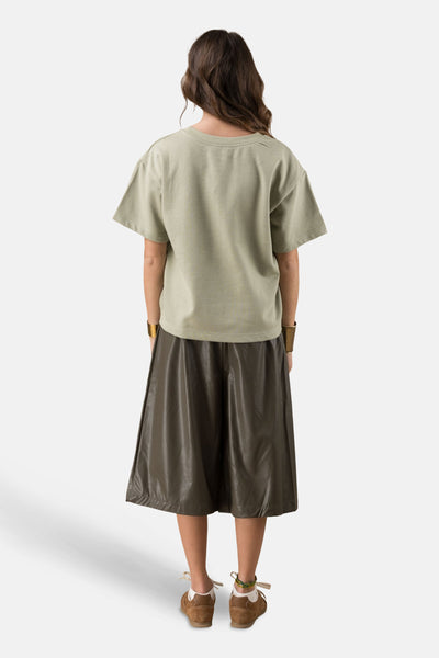 ADORE kniebroek, knielengte, gedragen met een olijfgroen T-shirt, gouden armbanden en bruin-witte sneakers.