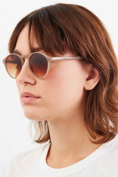 Lunettes de soleil beiges de KOMONO, monture ronde, verres dégradés marron, portées par une femme en t-shirt blanc.
