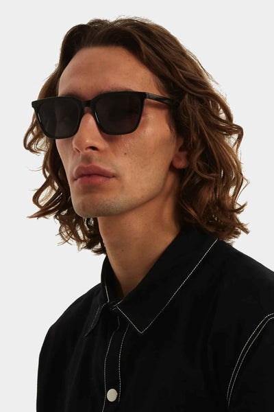 Lunettes de soleil noires de KOMONO, portées par un homme avec une chemise noire à coutures blanches.
