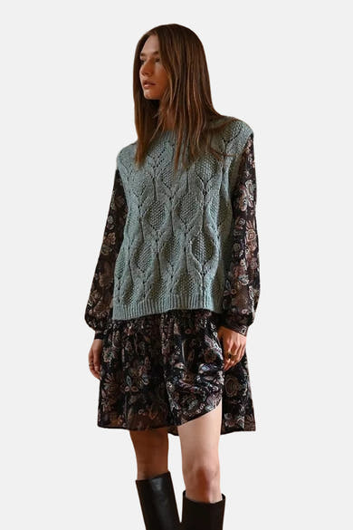 Blauwe gebreide tanktop van Molly Bracken, gedragen over een bloemenjurk met lange mouwen en zwarte laarzen.