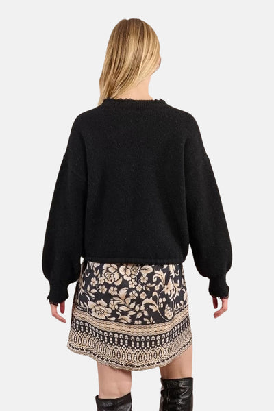 Pull à col rond noir de Molly Bracken, manches bouffantes, porté avec une jupe à motif floral et des bottes noires.
