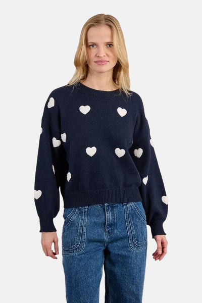 Pull bleu à col rond de Molly Bracken, orné de motifs de coeurs blancs et porté avec un jean.
