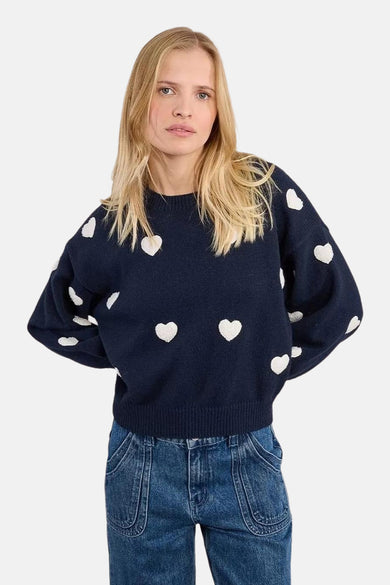 Pull à col rond bleu de Molly Bracken, avec motif de cœurs blancs, porté avec un jean bleu.
