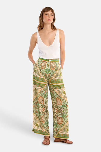 Pantalon large multicolore de Molly Bracken à motifs paisley verts, oranges et beiges, avec débardeur blanc et sandales.