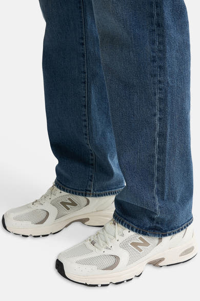 Mid blue denim jeans by LEWIS, wide-leg fit with faded wash and cuffed hem, shown with white mesh sneakers, side view.
