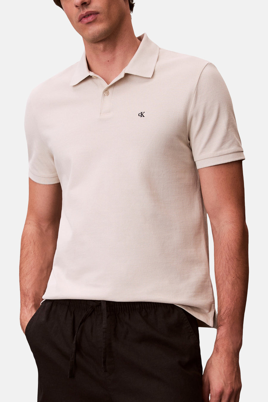 Polo à manches courtes écru de Calvin Klein, avec logo brodé sur la poitrine, porté avec un pantalon noir décontracté.

