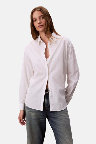Chemise blanche à manches longues de Calvin Klein, portée sur un soutien-gorge, avec un jean taille basse et une ceinture noire.

