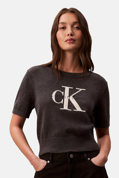 Grijs rondehals-trui van Calvin Klein, met wit logo, gedragen met een zwarte jas en broek.