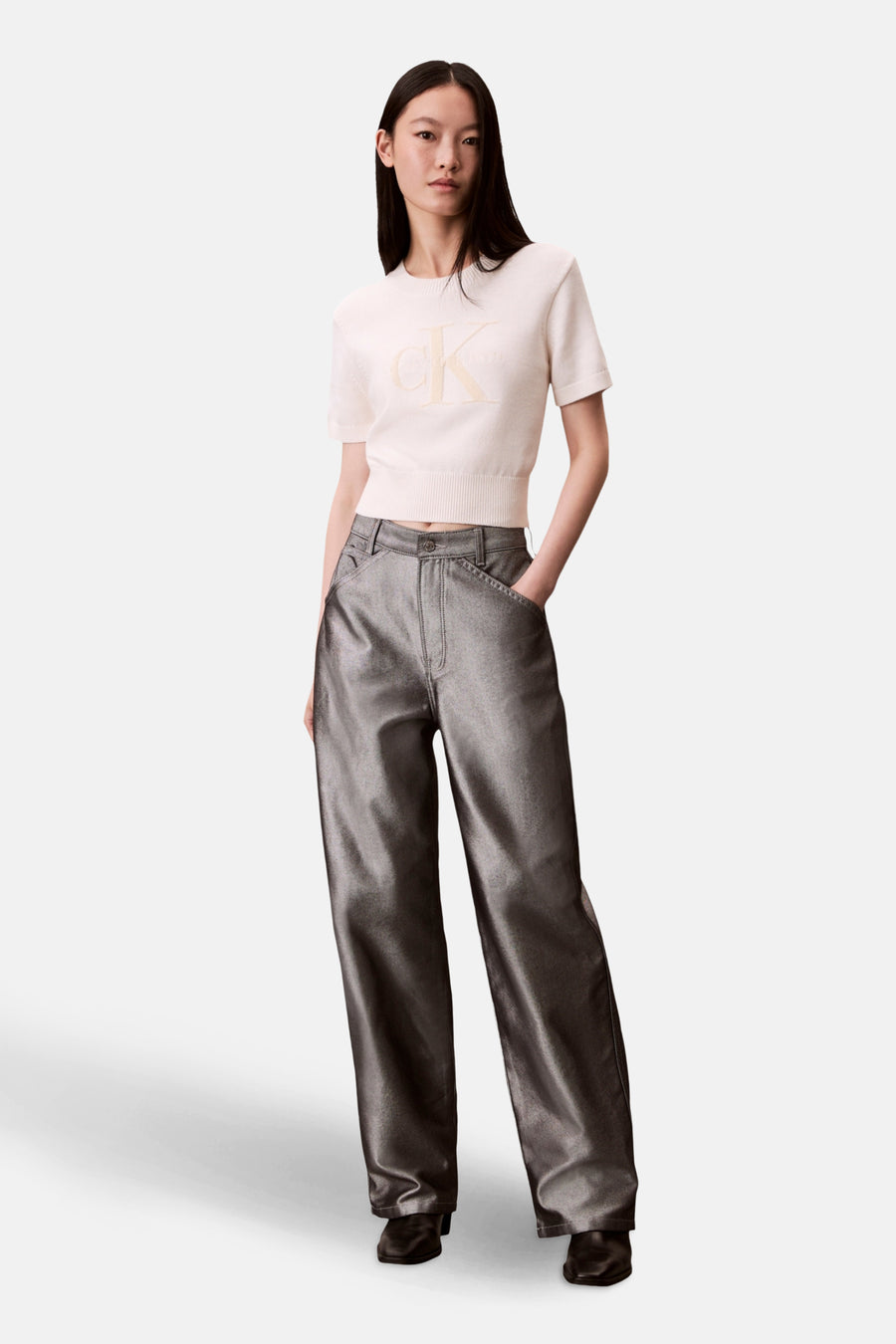 Pull à col rond écru de Calvin Klein, avec le logo sur le devant, porté avec un pantalon argenté.