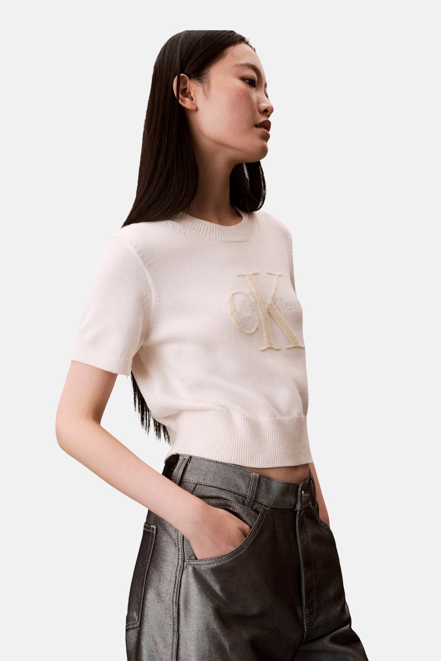 Pull écru à col rond de Calvin Klein, avec le logo brodé, porté avec un pantalon gris métallisé.

