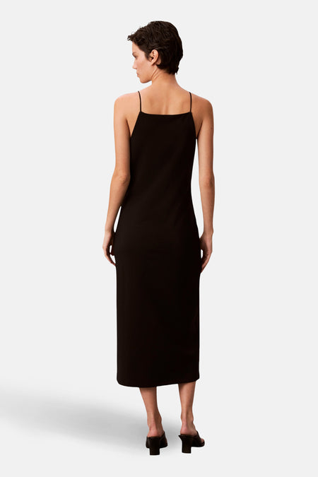 Robe noire simple de Calvin Klein, coupe midi avec bretelles fines et chaussures à talons.
