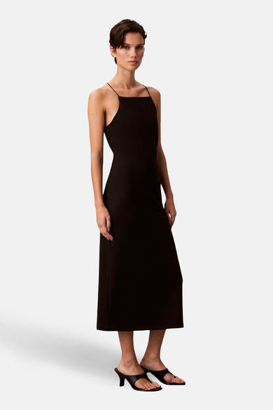 Robe noire moulante de Calvin Klein, mi-longue et à fines bretelles, portée avec des mules noires à talons.
