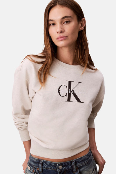 Sweat-shirt beige à col rond de Calvin Klein avec logo noir sur le devant, porté avec un jean bleu.
