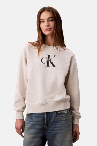 Sweat beige en coton mélangé de Calvin Klein, avec logo noir sur le devant et porté avec un jean bleu.
