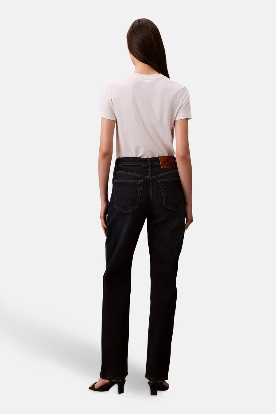 Rechte donkerblauwe spijkerbroek van Calvin Klein, gedragen met een wit T-shirt en zwarte sandalen, achterzicht.