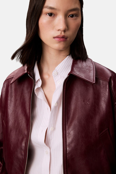 Veste courte bordeaux en cuir synthétique de Calvin Klein, avec fermeture éclair et rayures verticales sur la chemise portée en dessous.
