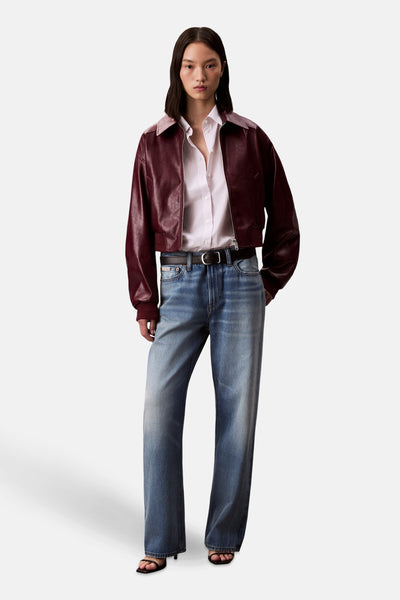 Veste courte bordeaux en cuir par Calvin Klein, portée avec une chemise rayée, un jean bleu et des sandales noires.