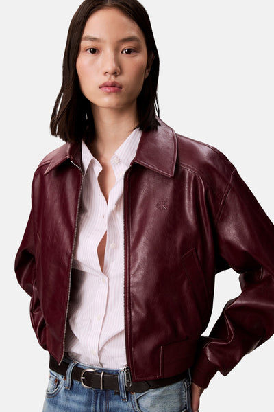 Veste courte bordeaux en cuir synthétique de Calvin Klein, portée sur une chemise rayée et un jean bleu.
