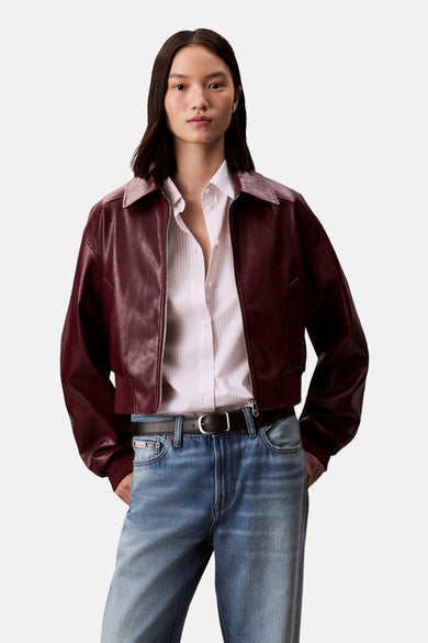 Veste courte bordeaux en cuir de Calvin Klein, portée sur une chemise rayée et un jean bleu classique avec une ceinture noire.

