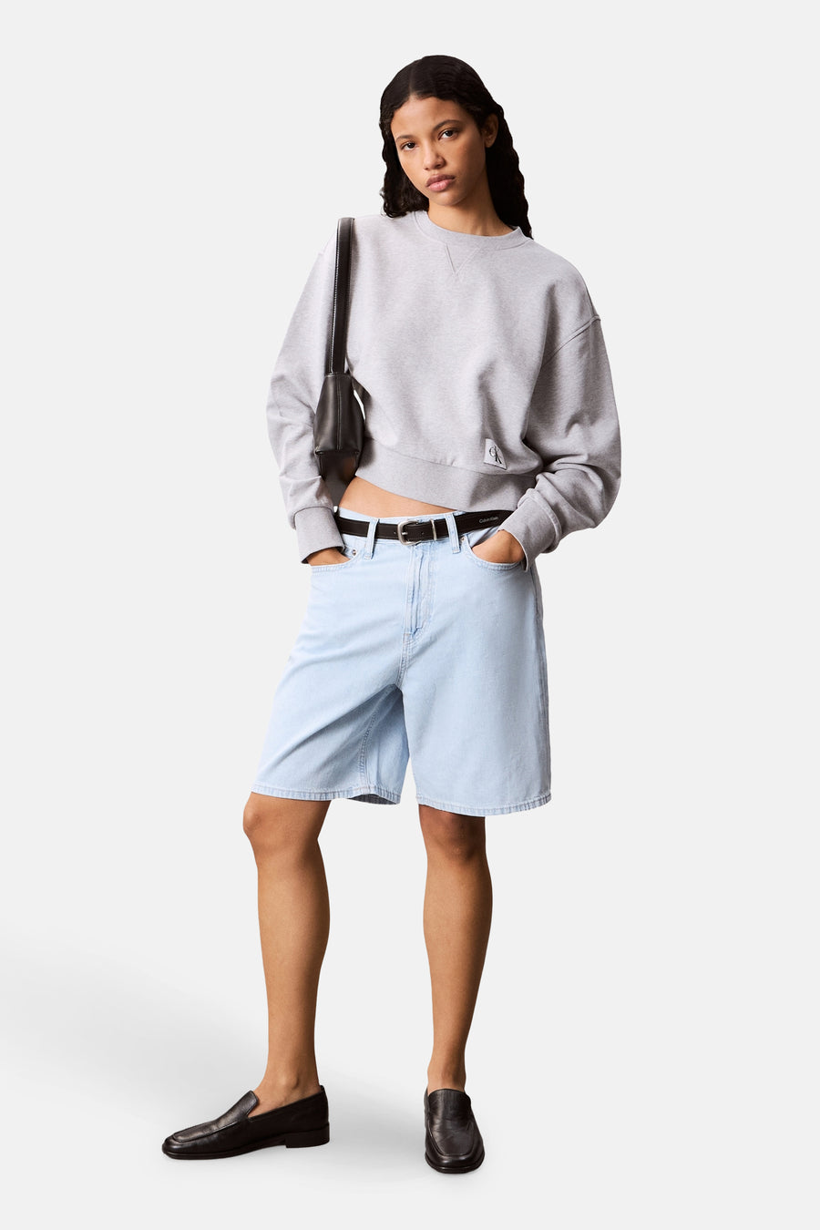Sweat col rond gris de Calvin Klein, porté avec un short en jean bleu clair, une ceinture et des mocassins noirs.

