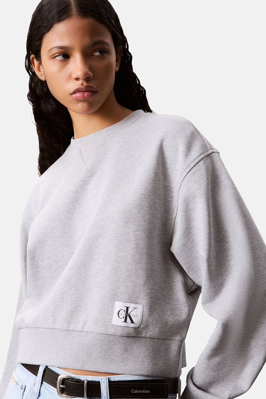 Sweat-shirt col rond gris chiné de Calvin Klein, avec logo sur le devant, porté avec une ceinture en cuir noir.
