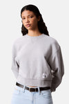 Sweat col rond gris de Calvin Klein, avec un logo sur le devant et porté avec un jean et une ceinture noirs.
