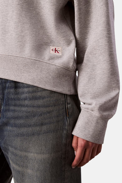 Sweat-shirt gris chiné par Calvin Klein, avec un logo carré rouge sur le côté, porté avec un jean bleu