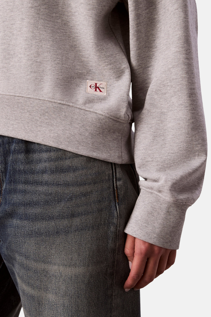 Sweat-shirt gris chiné par Calvin Klein, avec un logo carré rouge sur le côté, porté avec un jean bleu
