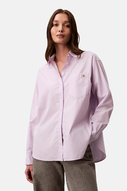 Chemise à manches longues violette de Calvin Klein, avec poche poitrine et boutons, portée avec un pantalon gris texturé.
