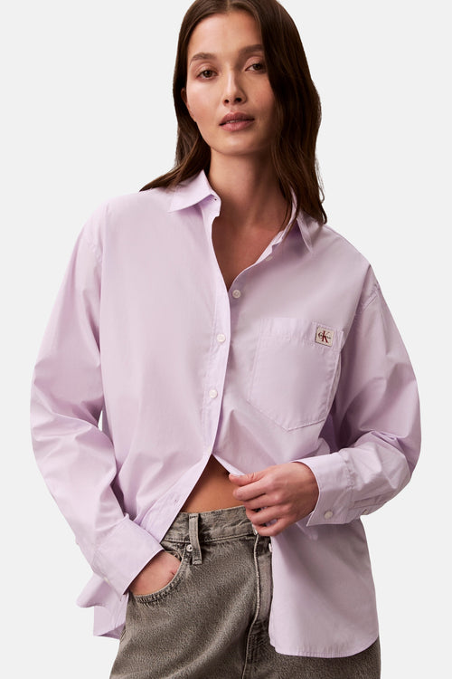 Chemise à manches longues violette de Calvin Klein, avec une poche poitrine et un logo discret, portée avec un jean taille haute.
