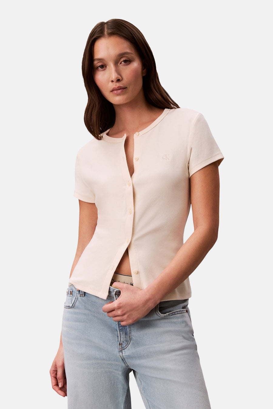 Ecru Calvin Klein vest open gedragen, met een olijfgroene trenchcoat, een verbleekte spijkerbroek en beige ballerina's met patroon.