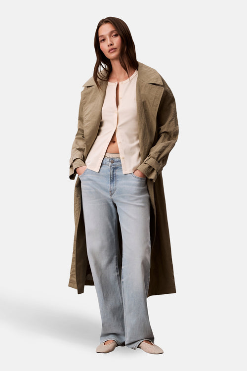 Gilet écru de Calvin Klein porté ouvert, avec un trench couleur olive, un jean délavé et des ballerines beiges à motifs.
