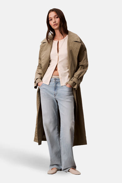 Gilet écru de Calvin Klein porté ouvert, avec un trench couleur olive, un jean délavé et des ballerines beiges à motifs.
