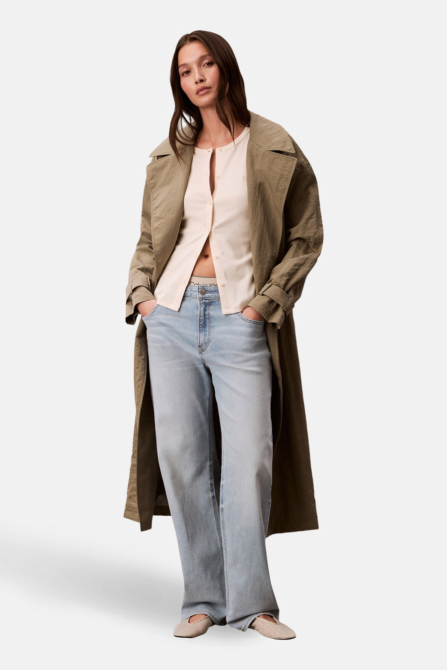 Gilet écru de Calvin Klein porté ouvert, avec un trench couleur olive, un jean délavé et des ballerines beiges à motifs.

