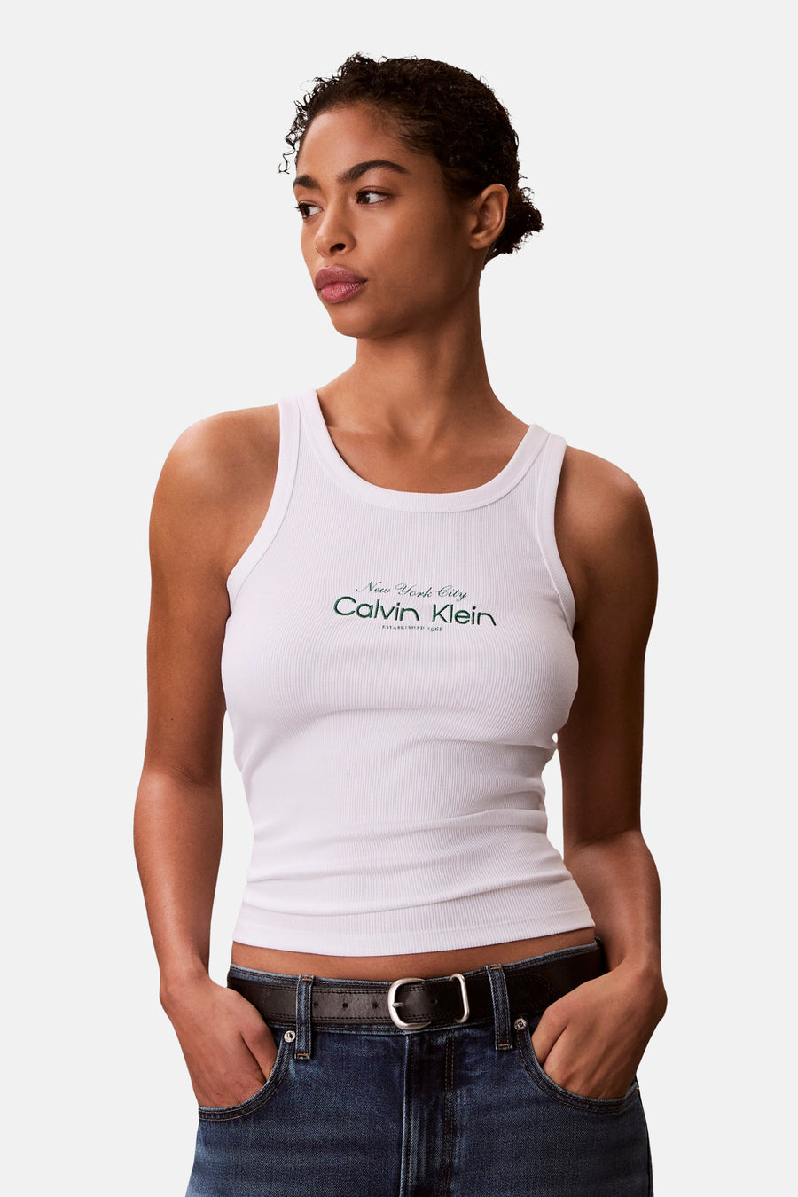 Witte geribde tanktop van Calvin Klein met groen logo, gedragen met een blauwe jeans en een zwarte riem.