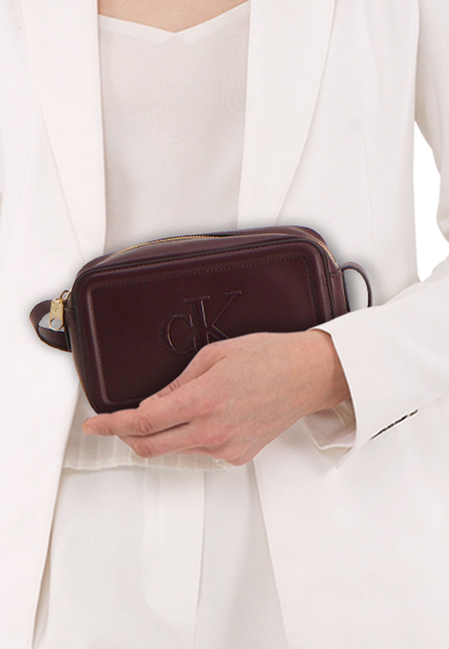 Sac à bandoulière bordeaux en cuir de Calvin Klein, porté à la main avec une veste blanche.
