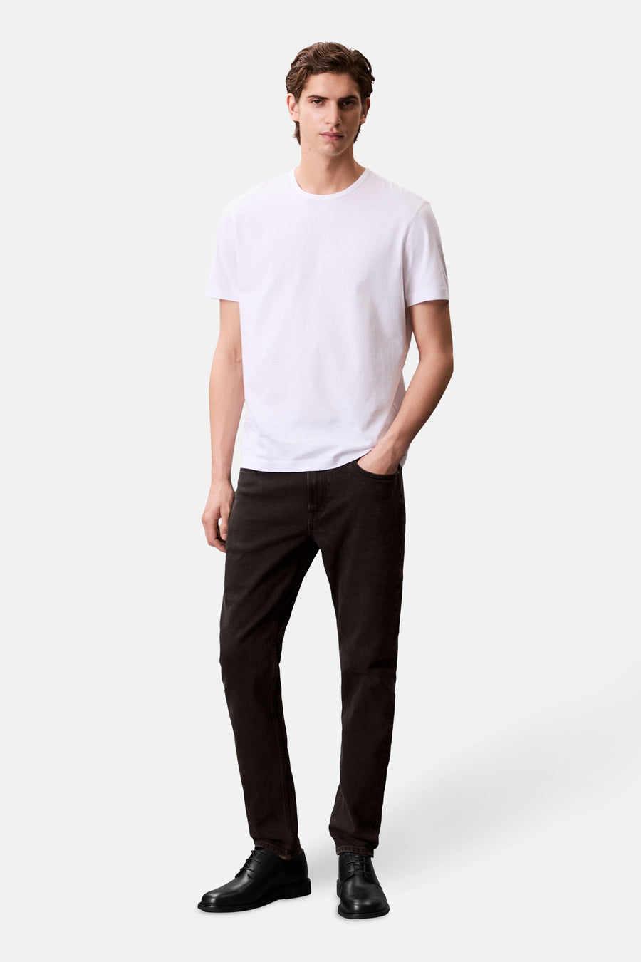 Jean tapered en denim gris foncé de Calvin Klein porté avec un t-shirt blanc uni et des chaussures noires.
