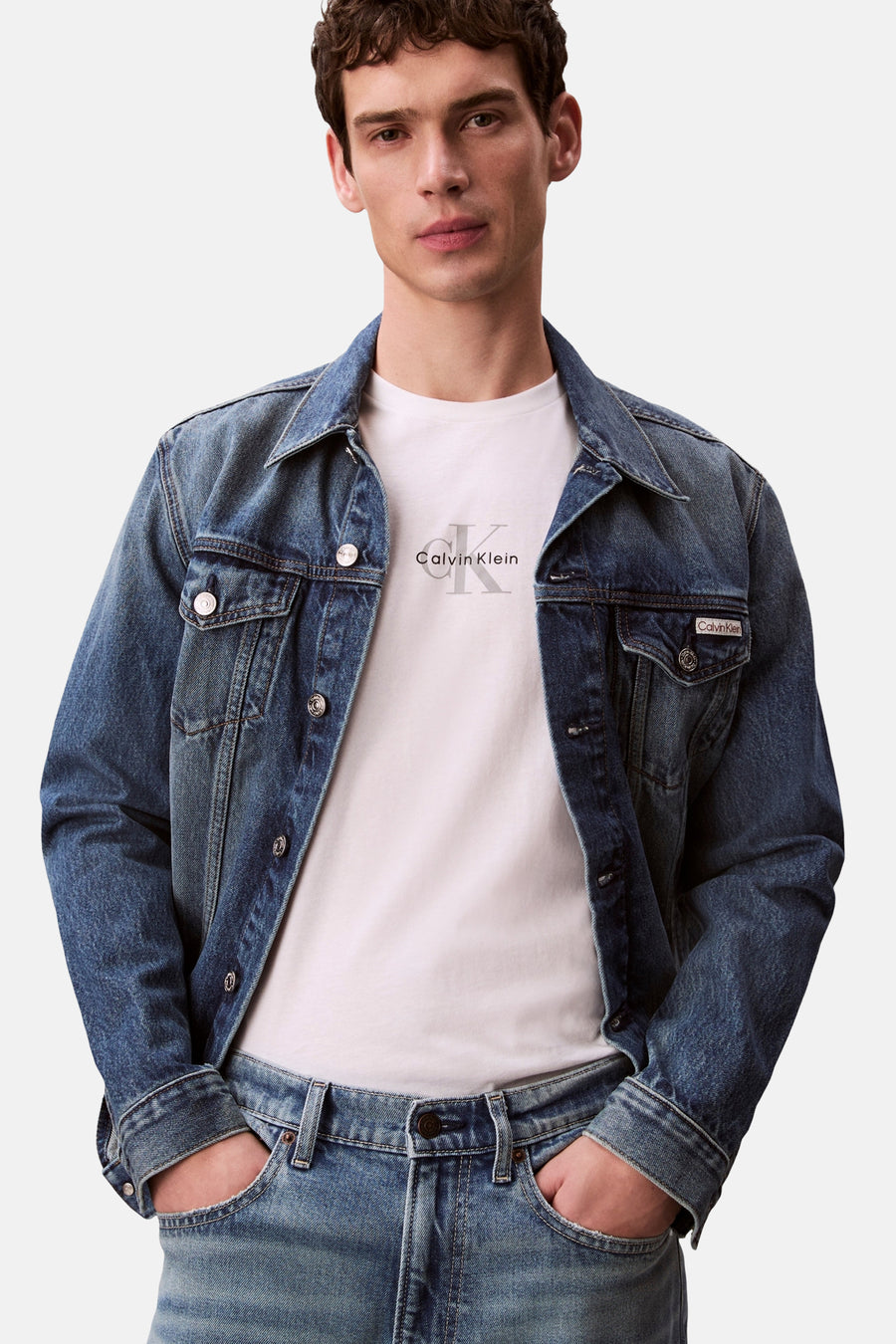 T-shirt à manches courtes blanc de Calvin Klein avec logo gris imprimé, porté sous une veste en jean.