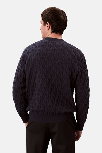 Sweat bleu col O de Calvin Klein, avec un motif texturé sur l'ensemble et porté avec un pantalon noir.
