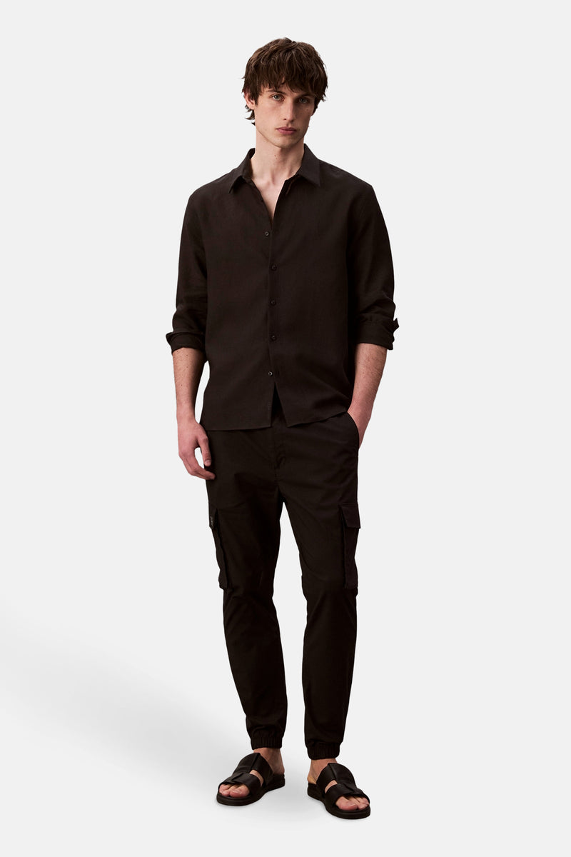 Pantalon cargo noir de Calvin Klein, avec poches latérales et coupe ajustée, porté avec une chemise boutonnée noire.