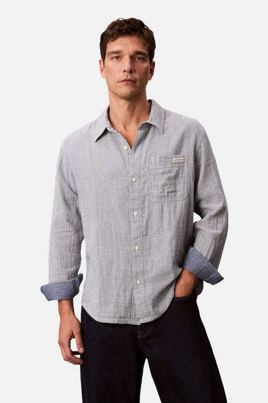 Chemise à manches longues bleue rayée de Calvin Klein, avec poche poitrine et manches retroussées sur un jean foncé.
