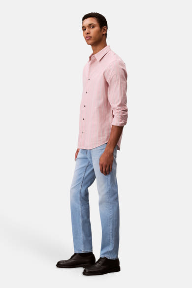 Slim jeans in lichtblauw denim van Calvin Klein, gedragen met een roze gestreept overhemd en zwarte schoenen, zijwaarts gezien.