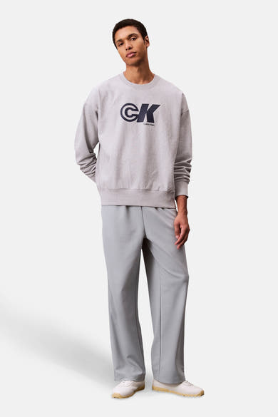 Sweat-shirt gris à col rond de Calvin Klein, avec le logo de la marque en bleu marine, porté avec un pantalon gris.

