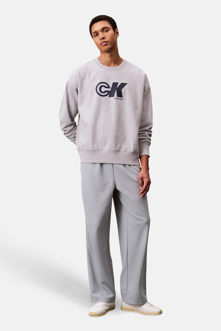 Sweat-shirt gris à col rond de Calvin Klein, avec le logo de la marque en bleu marine, porté avec un pantalon gris.
